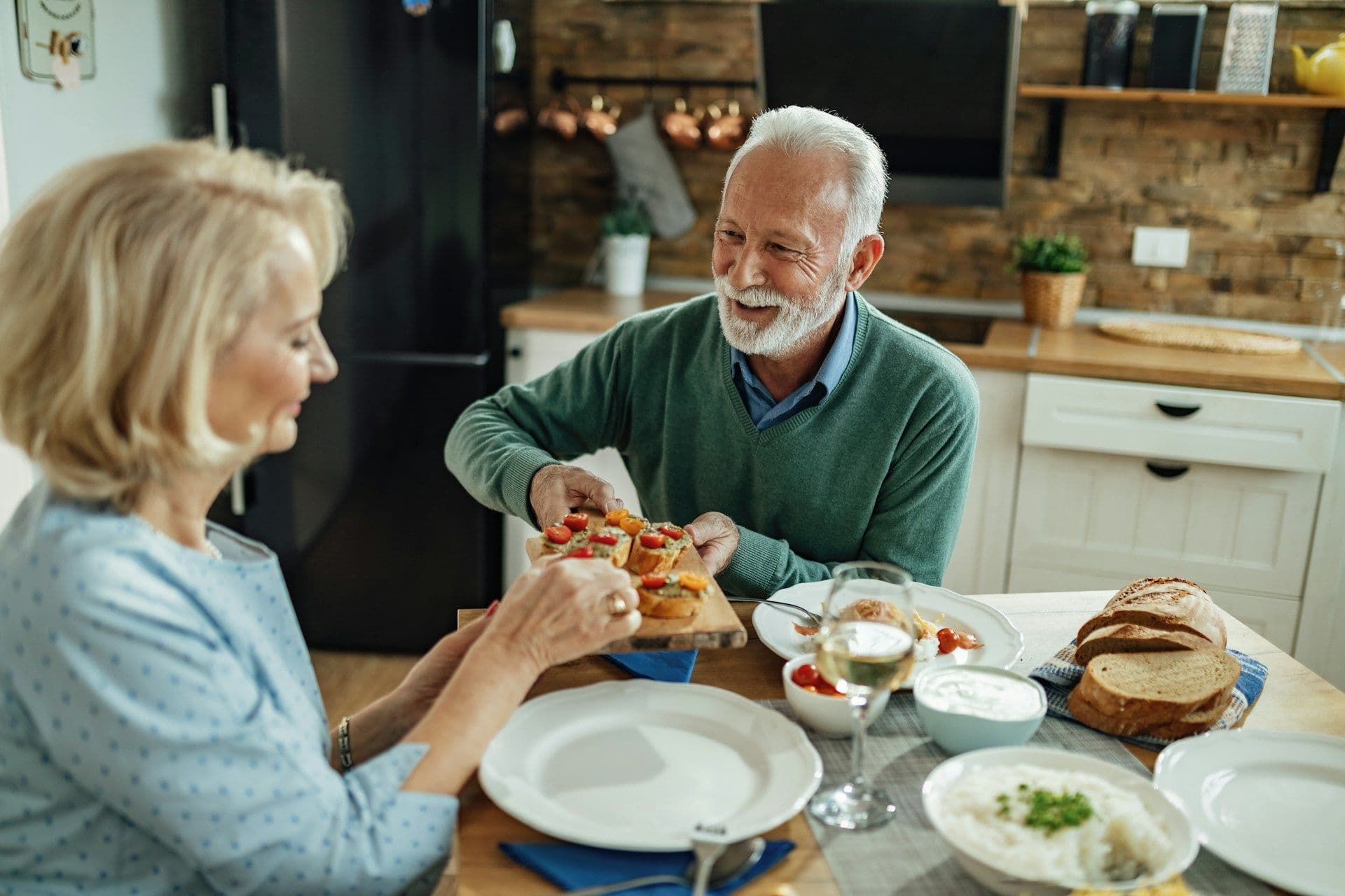 An older couple laughing as they share a board of snacks at the kitchen table